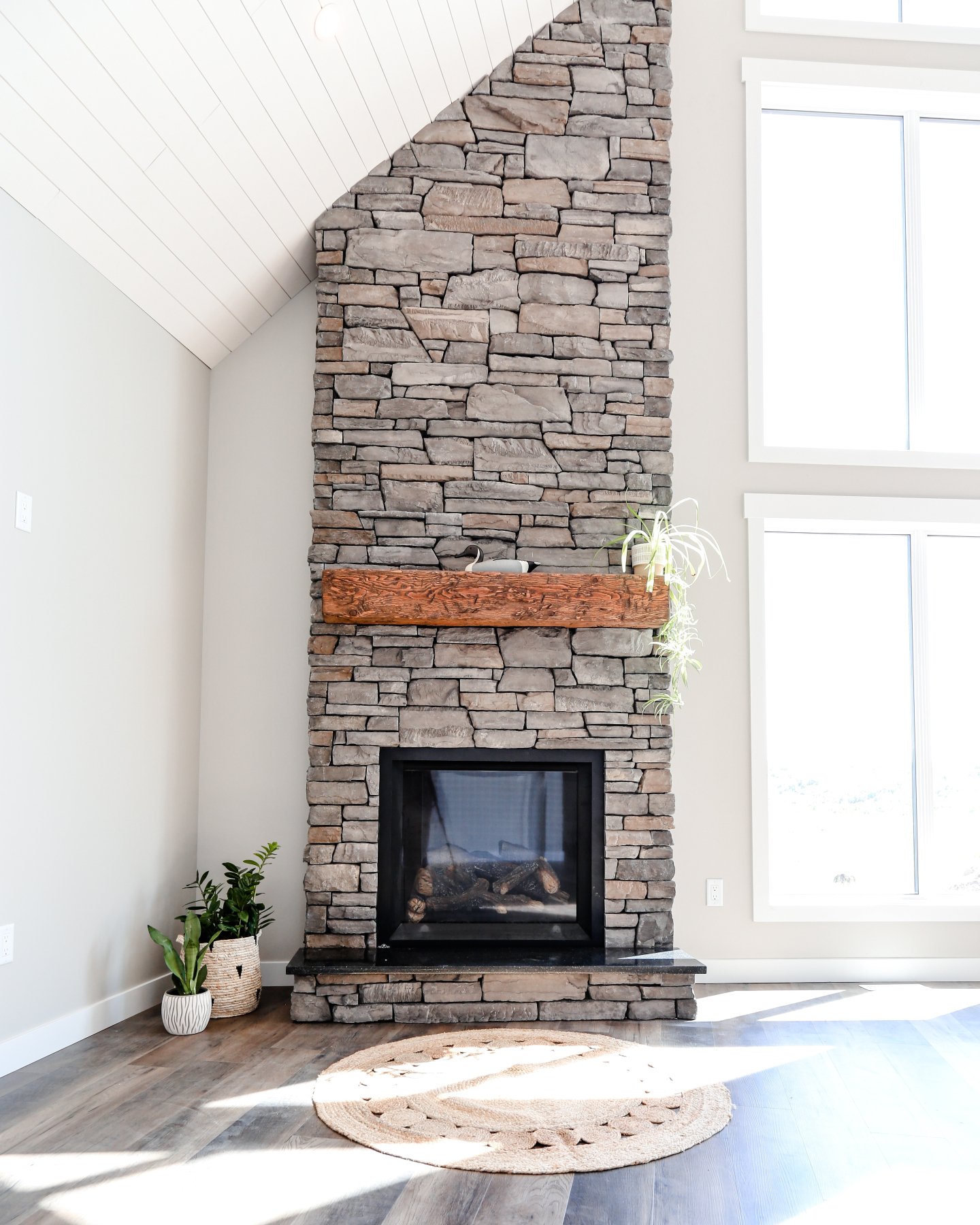 A tall, slender fireplace column made of mixed-size grey and brown rectangular-cut stone, with a rustic live-edge wood mantel, and a square black firebox, set beneath a vaulted ceiling.