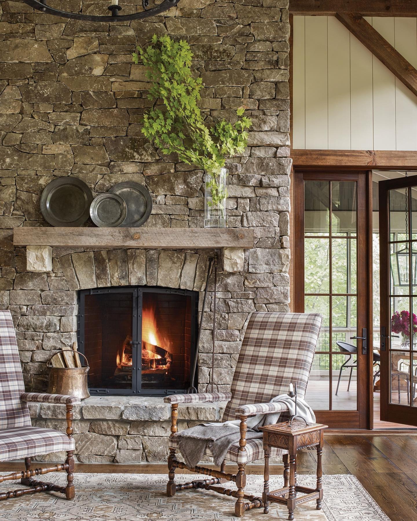 A tall, rustic stone fireplace with a thick wood beam mantel and a glowing fire, flanked by two tall, plaid wingback chairs in a room with timber accents and French doors.