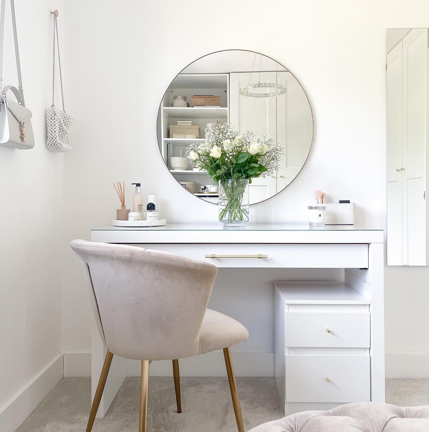 A white, modern dressing table with a glass top, a large circular mirror, and two storage drawers, paired with a beige velvet chair.