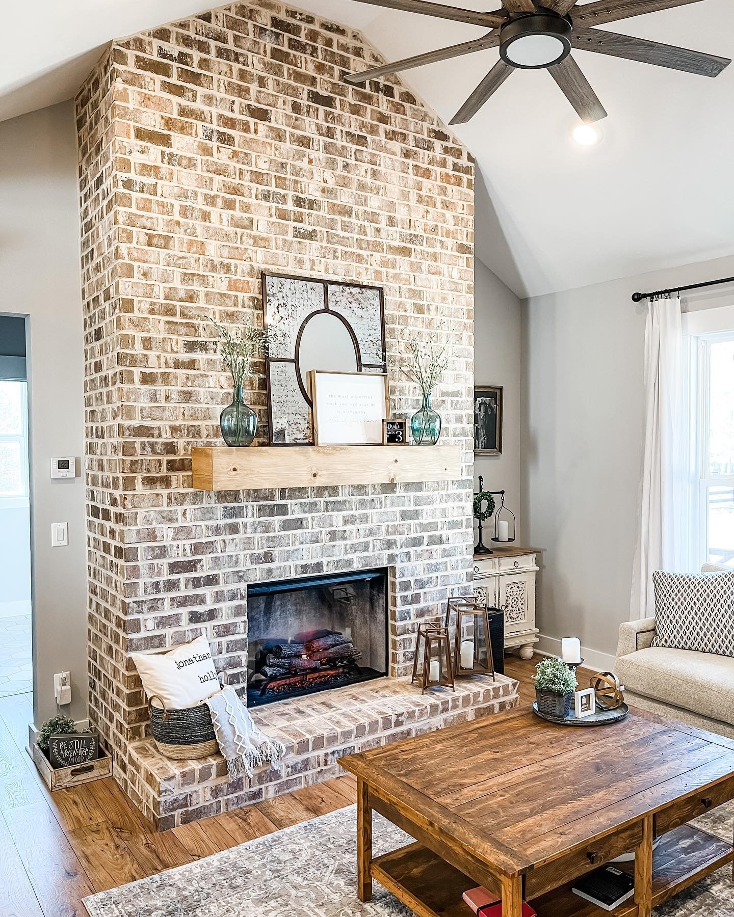 A tall, floor-to-ceiling brick fireplace with a distressed light finish. A thick wood mantel holds a mirror and vases, and the hearth is decorated with lanterns.
