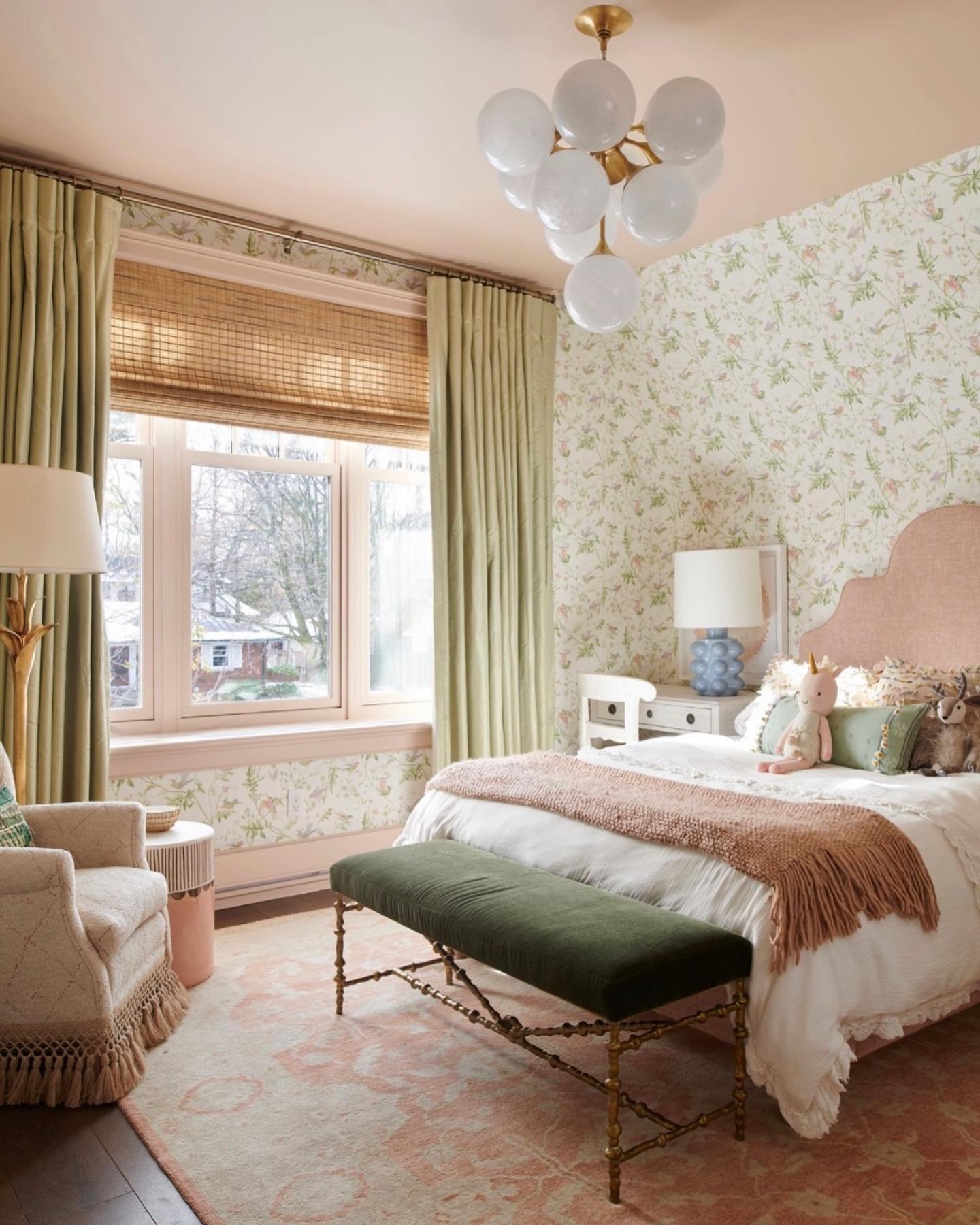 A playful bedroom featuring floral wallpaper, a blush pink upholstered headboard, sage green drapes, and a dark green velvet bench.