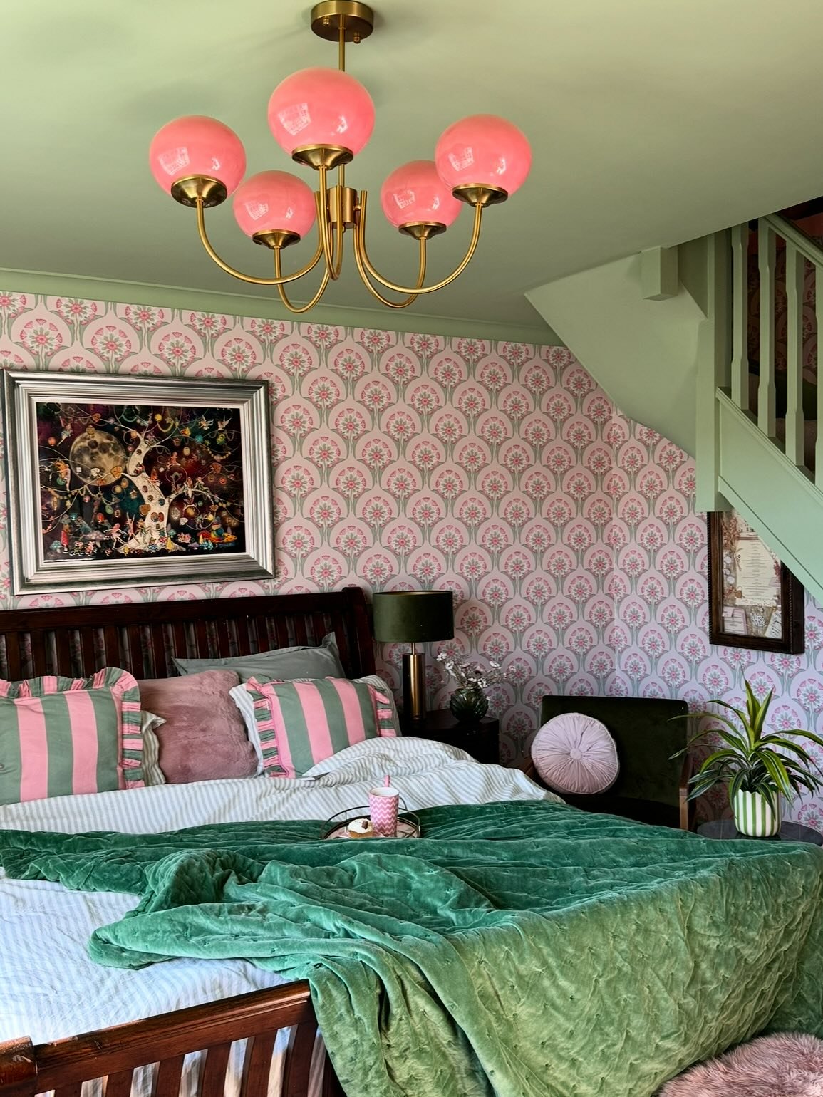 A vibrant bedroom featuring light green ceiling and woodwork, a pink and green patterned wallpaper, a dark wooden bed covered with a crushed green velvet throw, and a pink globe chandelier.