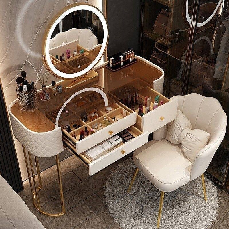 A luxurious cream and white dressing table with gold legs, a clear glass top revealing internal compartments, and an illuminated round mirror, paired with a quilted cream chair.