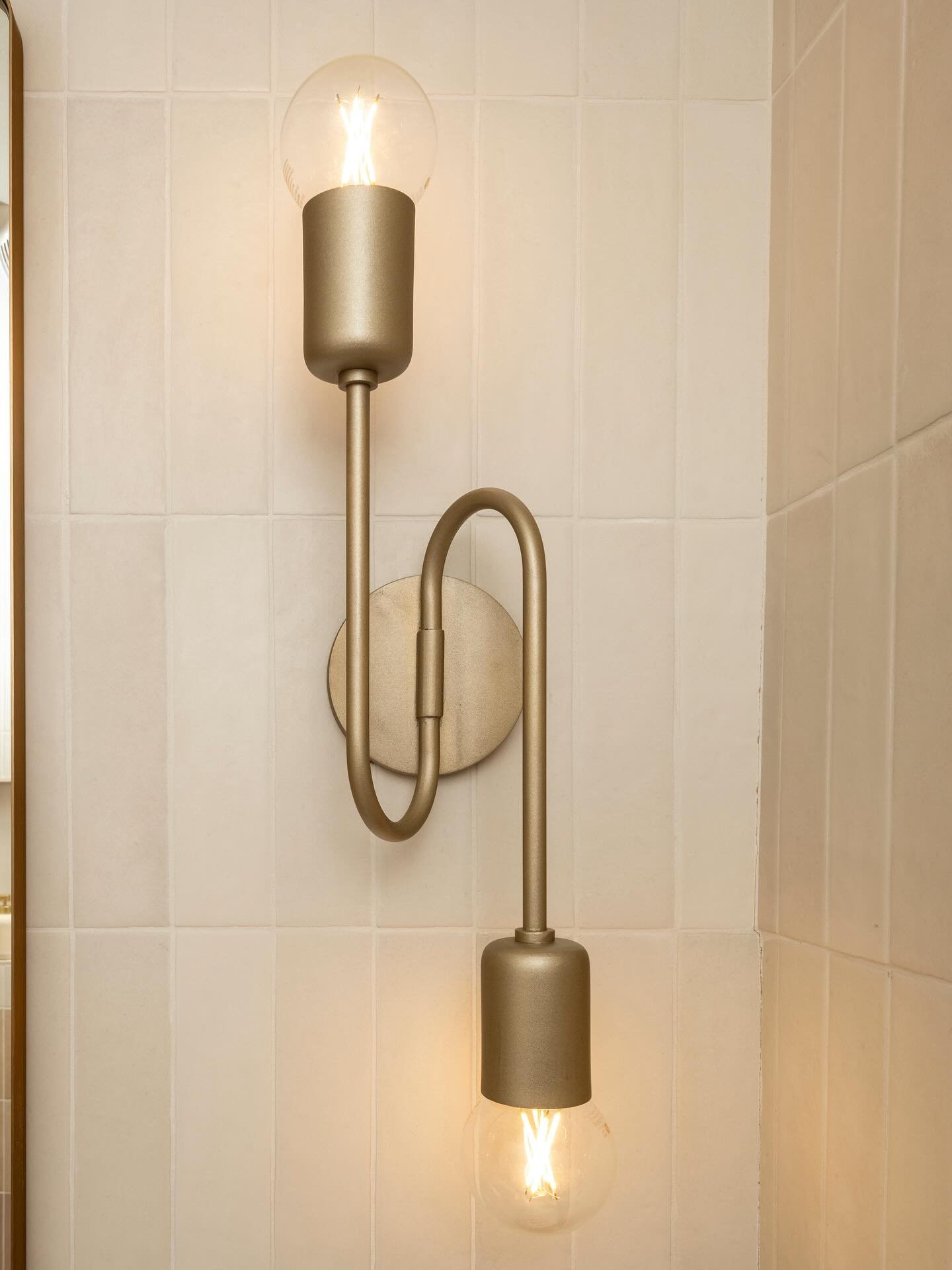 A brushed gold, s-shaped wall sconce with exposed filament bulbs on a tiled bathroom wall.