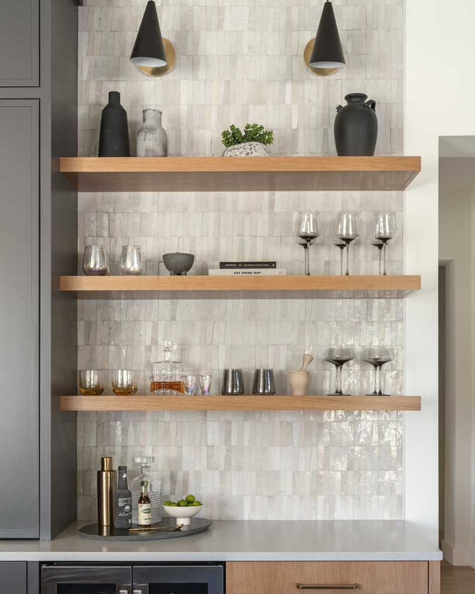 Three light wood floating shelves used for bar glassware against a textured tile backsplash.