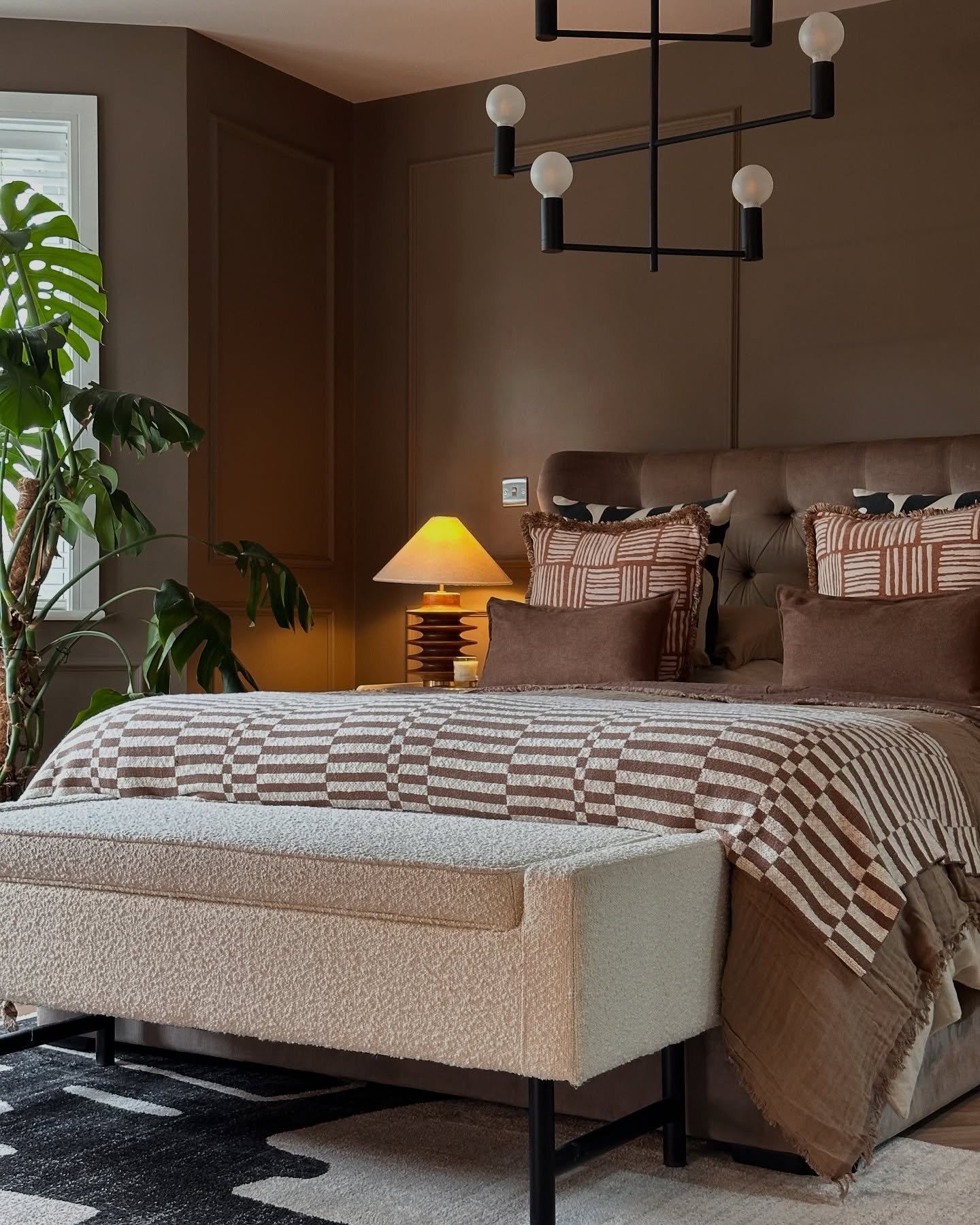 Brown bedroom with wall paneling, patterned bedding, a white boucle bench, and a large monstera plant