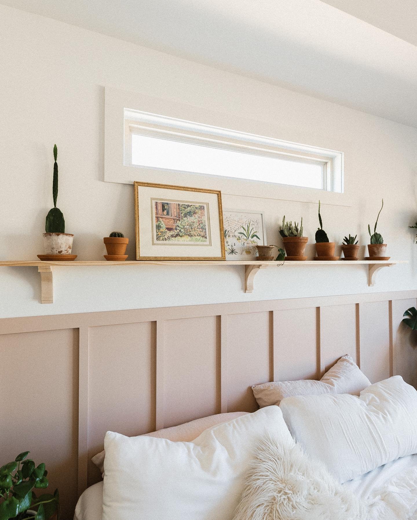 Long wooden shelf above pink paneling filled with cacti and art.