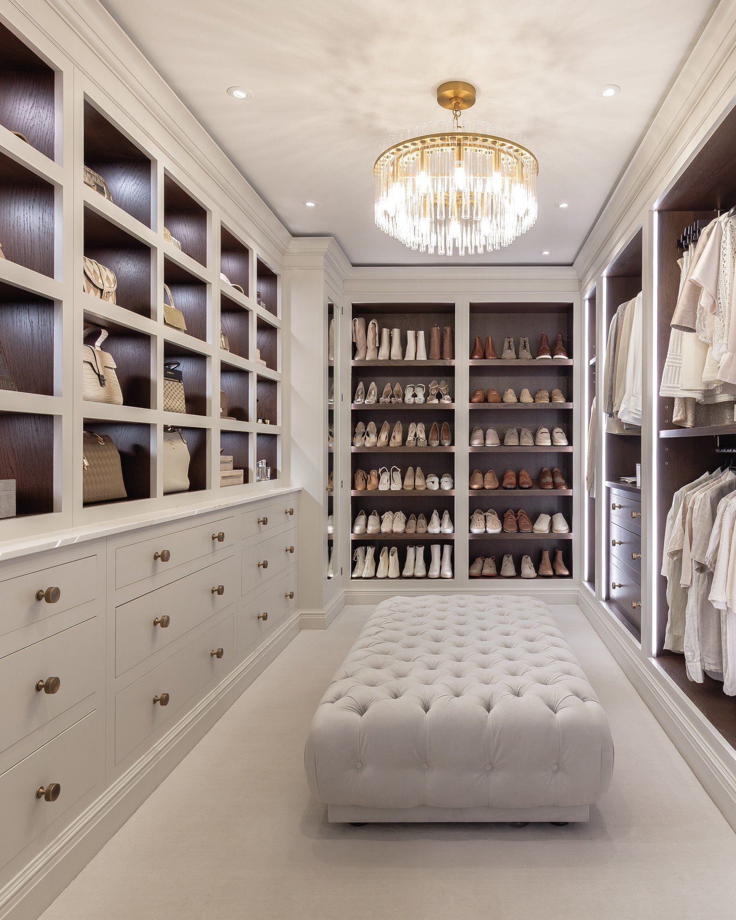 A neutral-toned walk-in closet featuring floor-to-ceiling shelving, a large tufted ottoman, and a modern gold and glass rod chandelier.