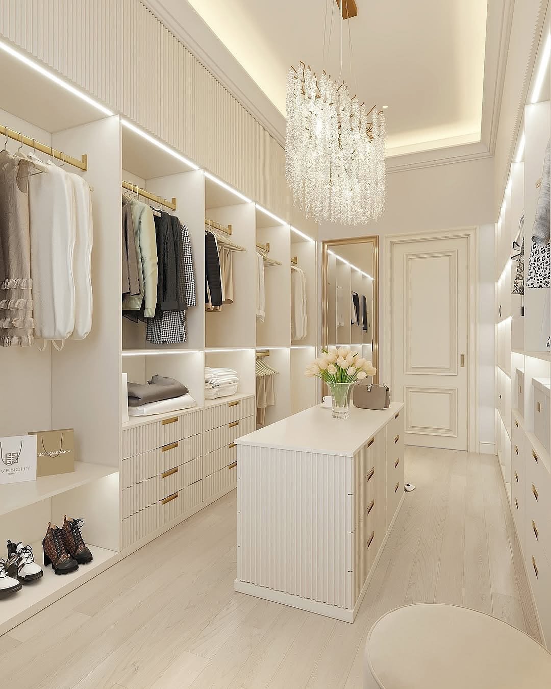 A bright, modern walk-in closet in white and cream tones featuring a textured central island, open shelving, and a waterfall-style crystal chandelier.