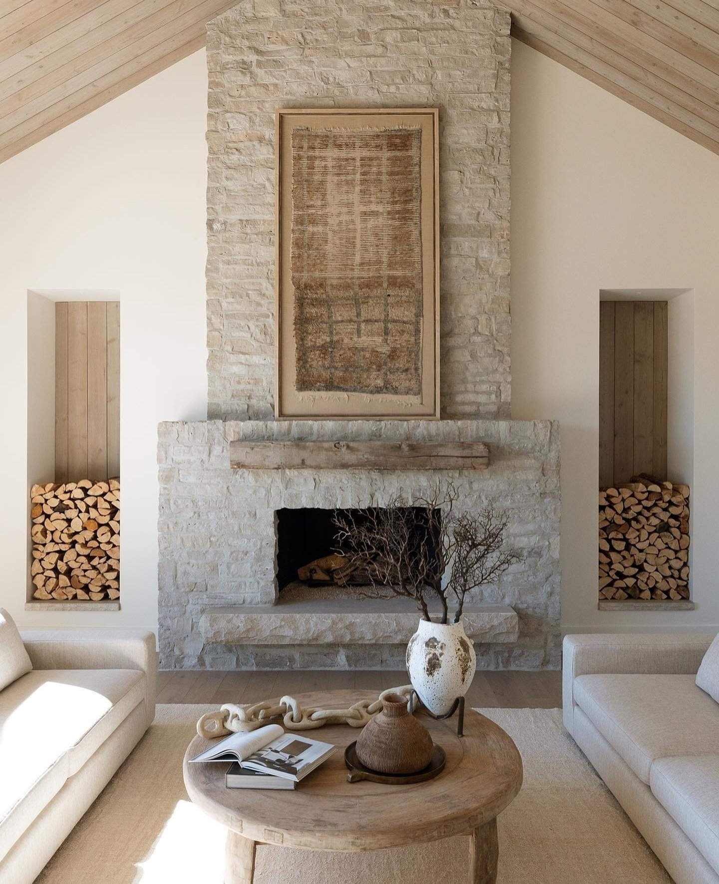 A symmetrical, vaulted-ceiling living room featuring a tall, light-colored limestone fireplace with a rough-hewn wood mantel and flanked by recessed niches for firewood storage.