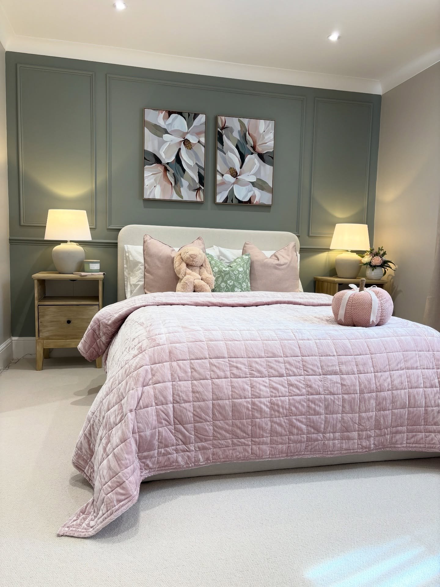 A bedroom featuring a paneled feature wall painted in a dusty sage green, a large bed covered with a blush pink quilted velvet blanket, and two floral prints above the headboard.