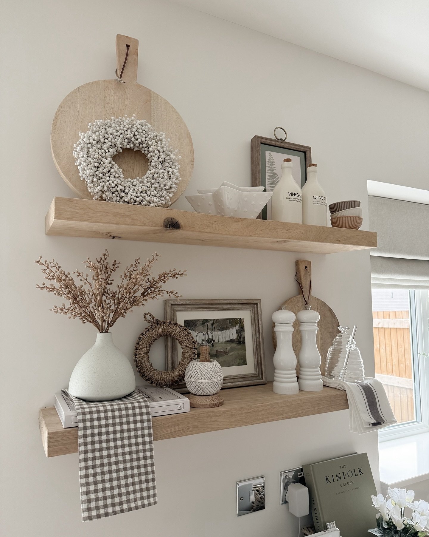 Two thick light wood floating shelves styled with neutral farmhouse decor, including cutting boards and white vases.