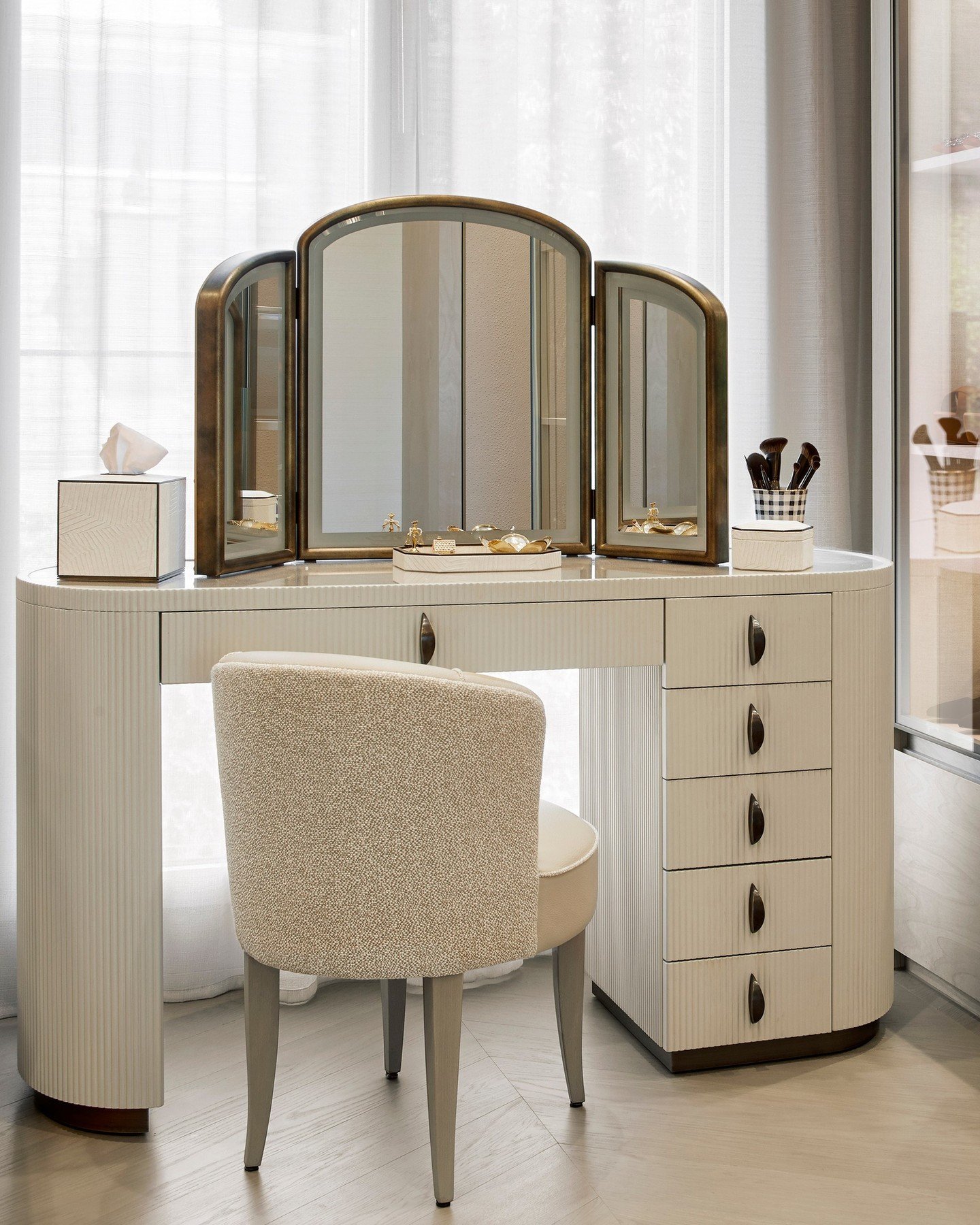 A cream-colored, rounded dressing table with vertical fluting, five drawers on the right, and a vintage-style bronze framed triptych mirror, paired with a textured cream chair.