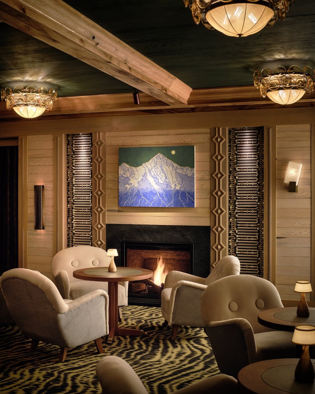 A dimly lit, wood-paneled room featuring a black marble fireplace surround, a fire burning within, and upholstered lounge chairs.