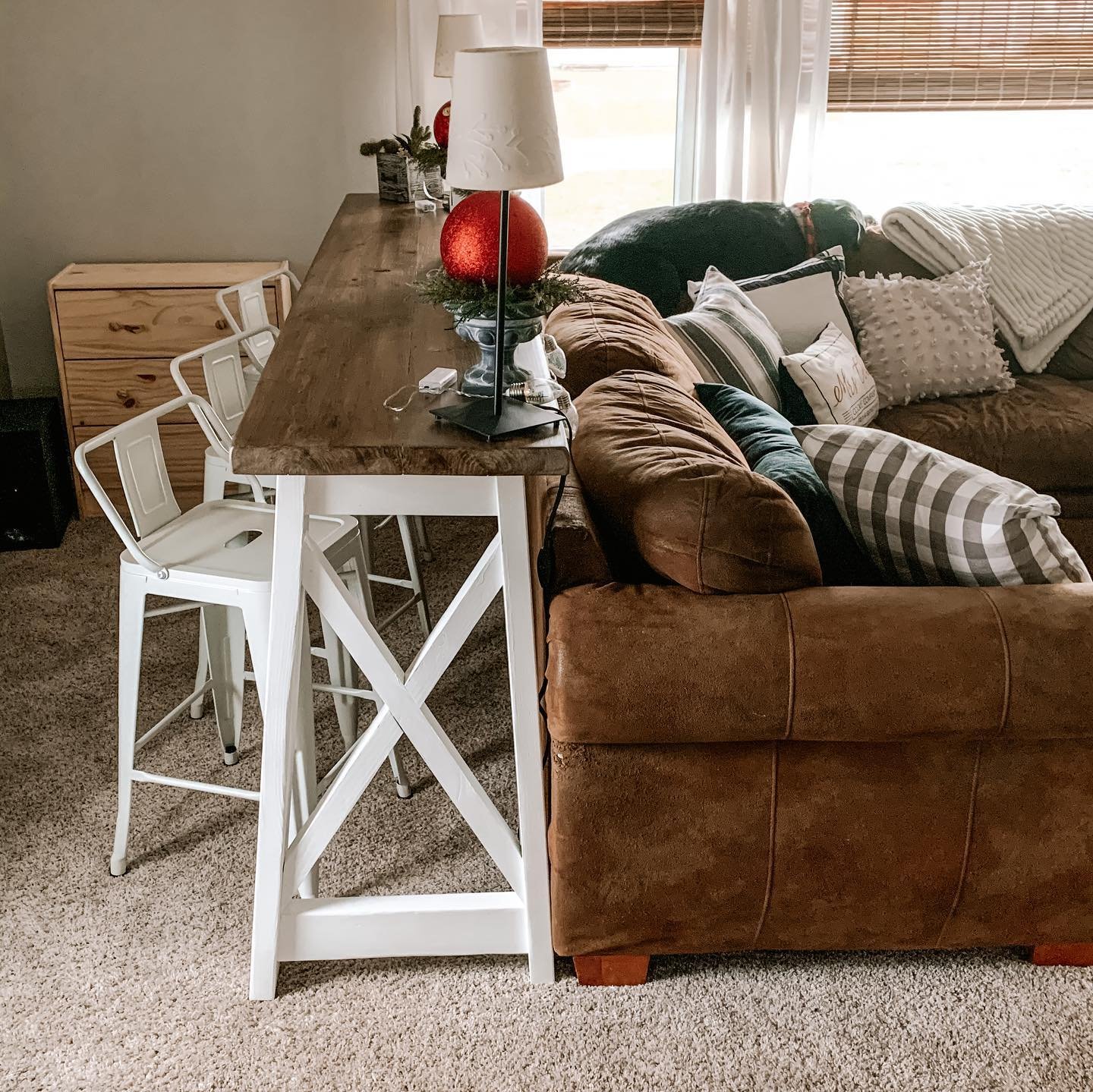 High top wooden table with white legs and stools behind brown couch