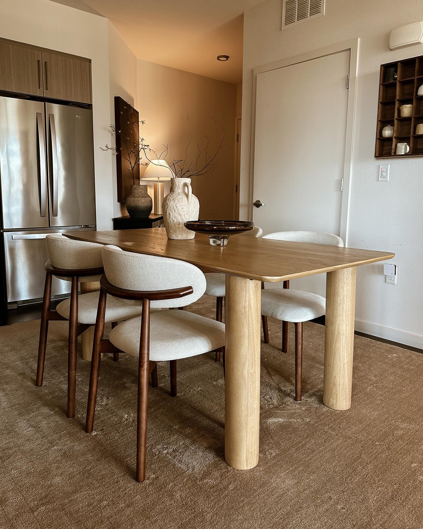Light wood dining table with thick round legs and curved white chairs near kitchen