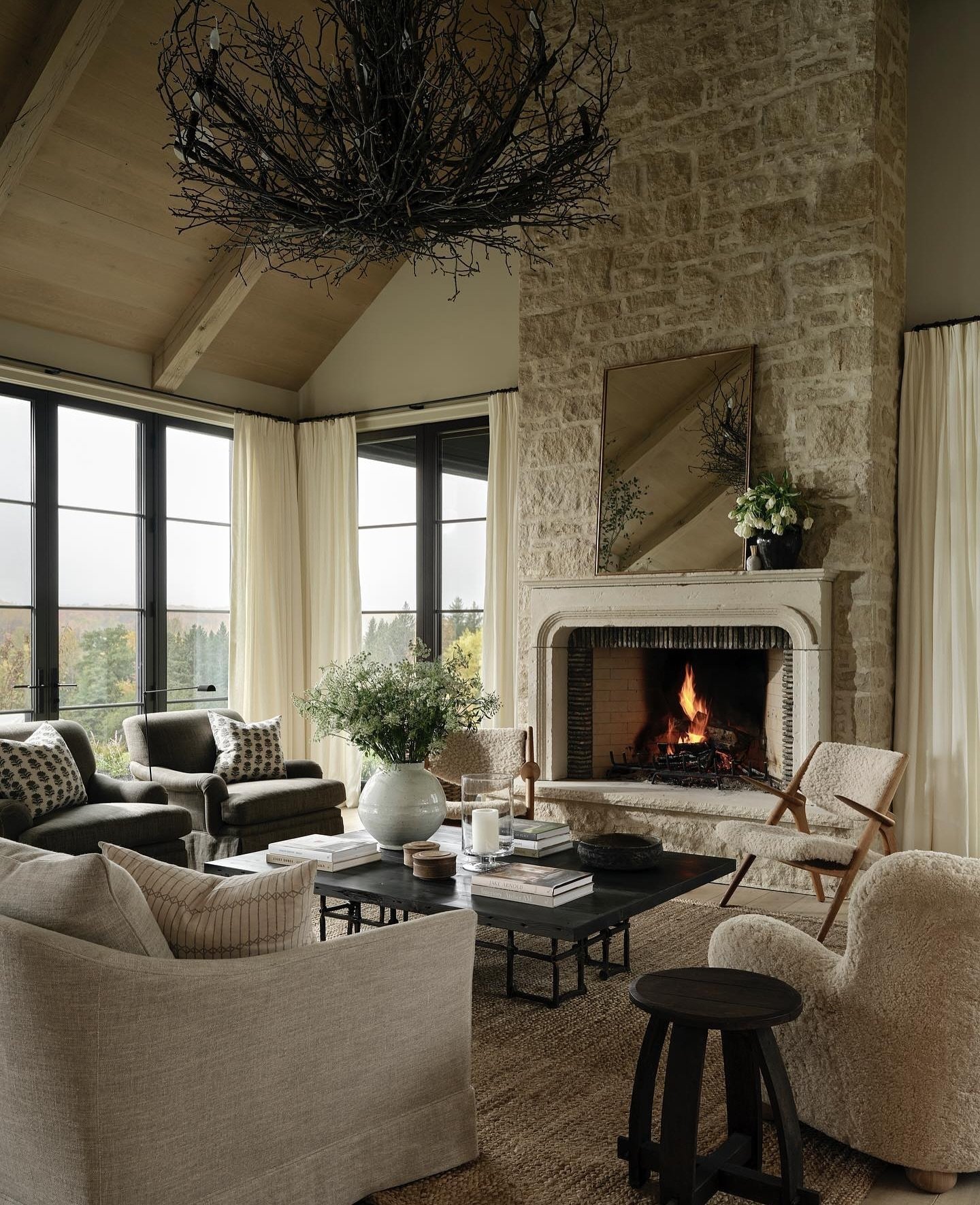 A tall, textured stone fireplace with a contrasting smooth white stone surround and mantel, featuring a cozy fire, set in a luxurious living room with large black-framed windows overlooking a landscape.