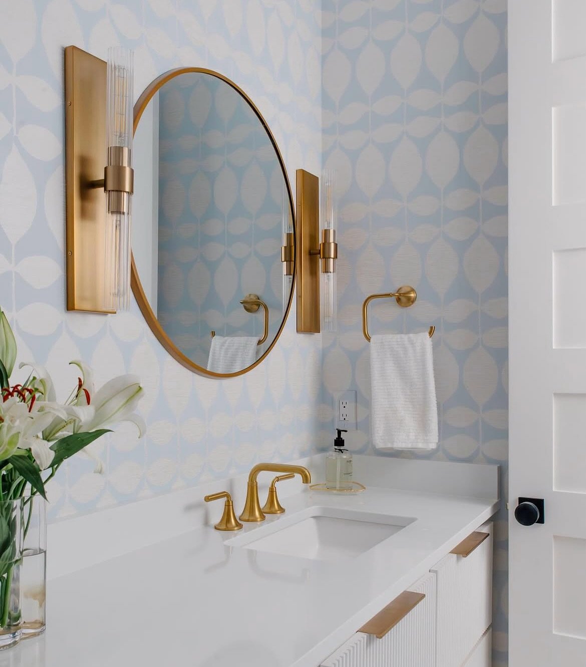 Vertical cylindrical glass and brass sconces framing a round gold mirror over a white vanity, set against blue and white patterned wallpaper.