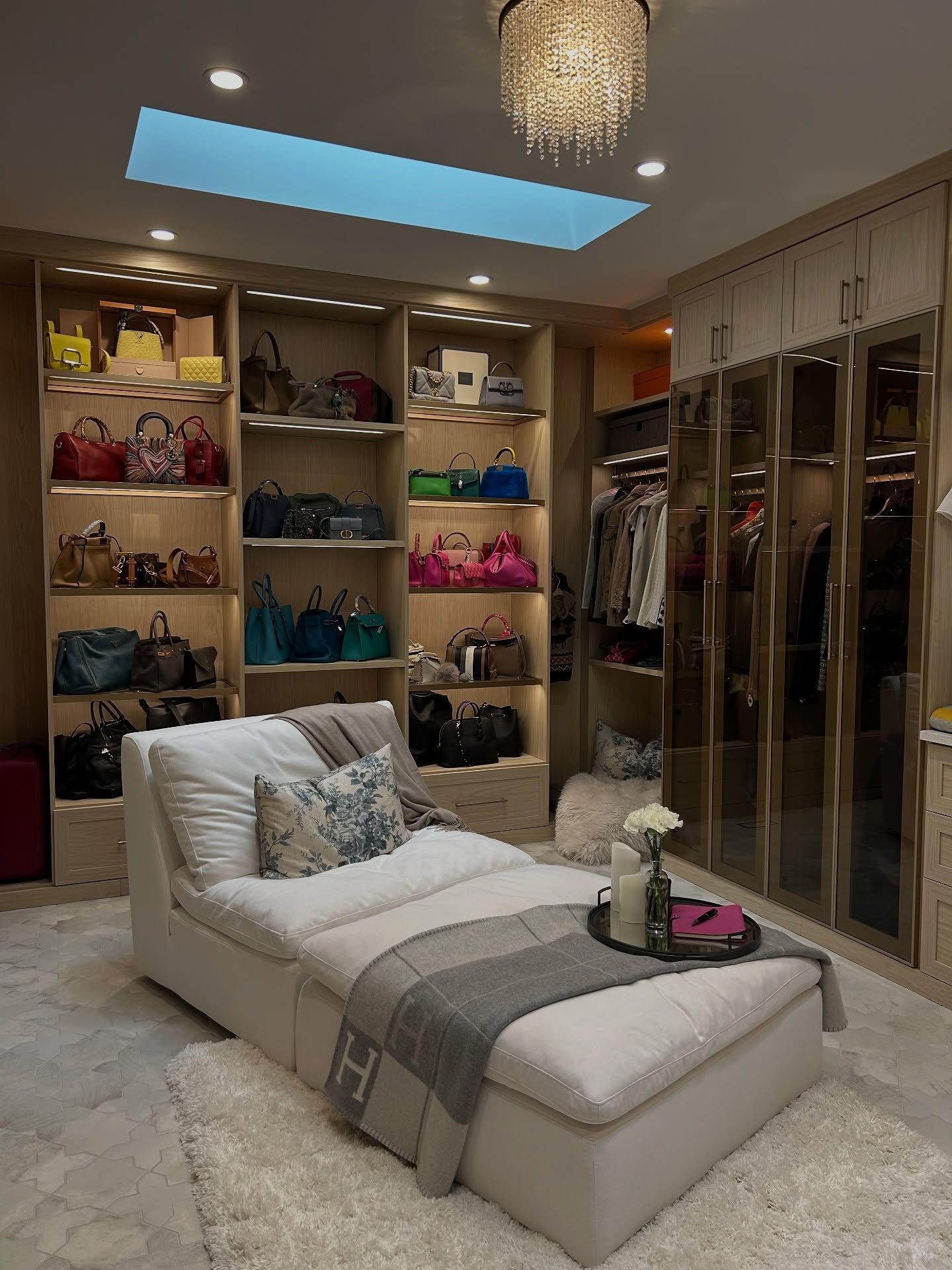 A spacious walk-in closet with light wood shelving dedicated to colorful designer handbags, featuring a large white chaise lounge and glass-fronted wardrobes.