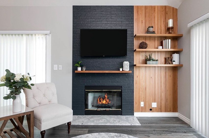 A contemporary living room wall unit featuring a central column of dark blue painted brick with a TV mounted above a black fireplace insert. To the right are floating wood shelves against a light wood paneled wall.