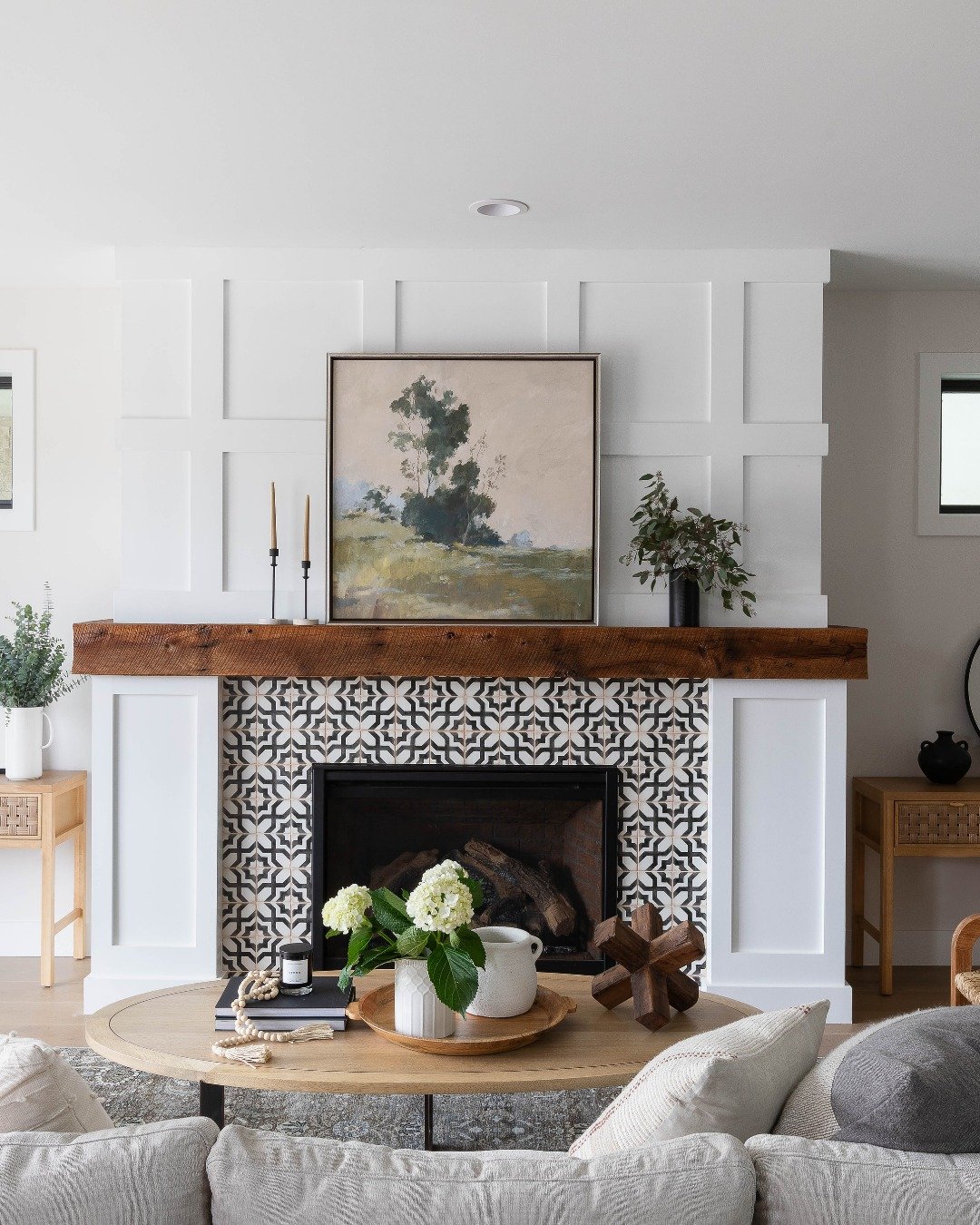 A fireplace with a rustic wood mantel and white paneling, featuring a surround tiled with intricate black and white patterned cement tiles.