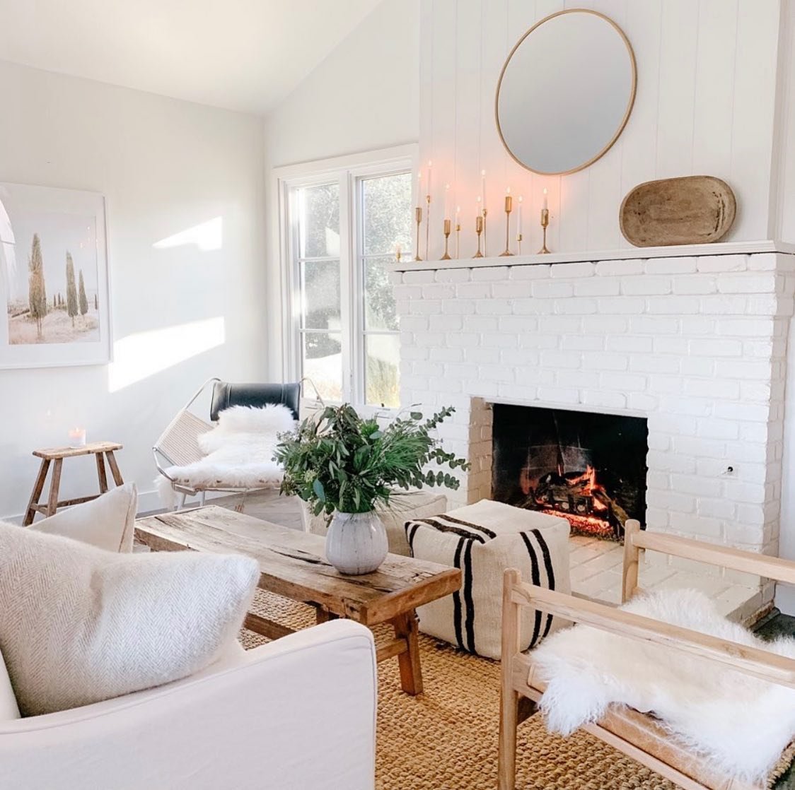 A white brick fireplace with a classic mantel, decorated with brass candlesticks and a round mirror. The fireplace sits beneath a wall with vertical white wood paneling.