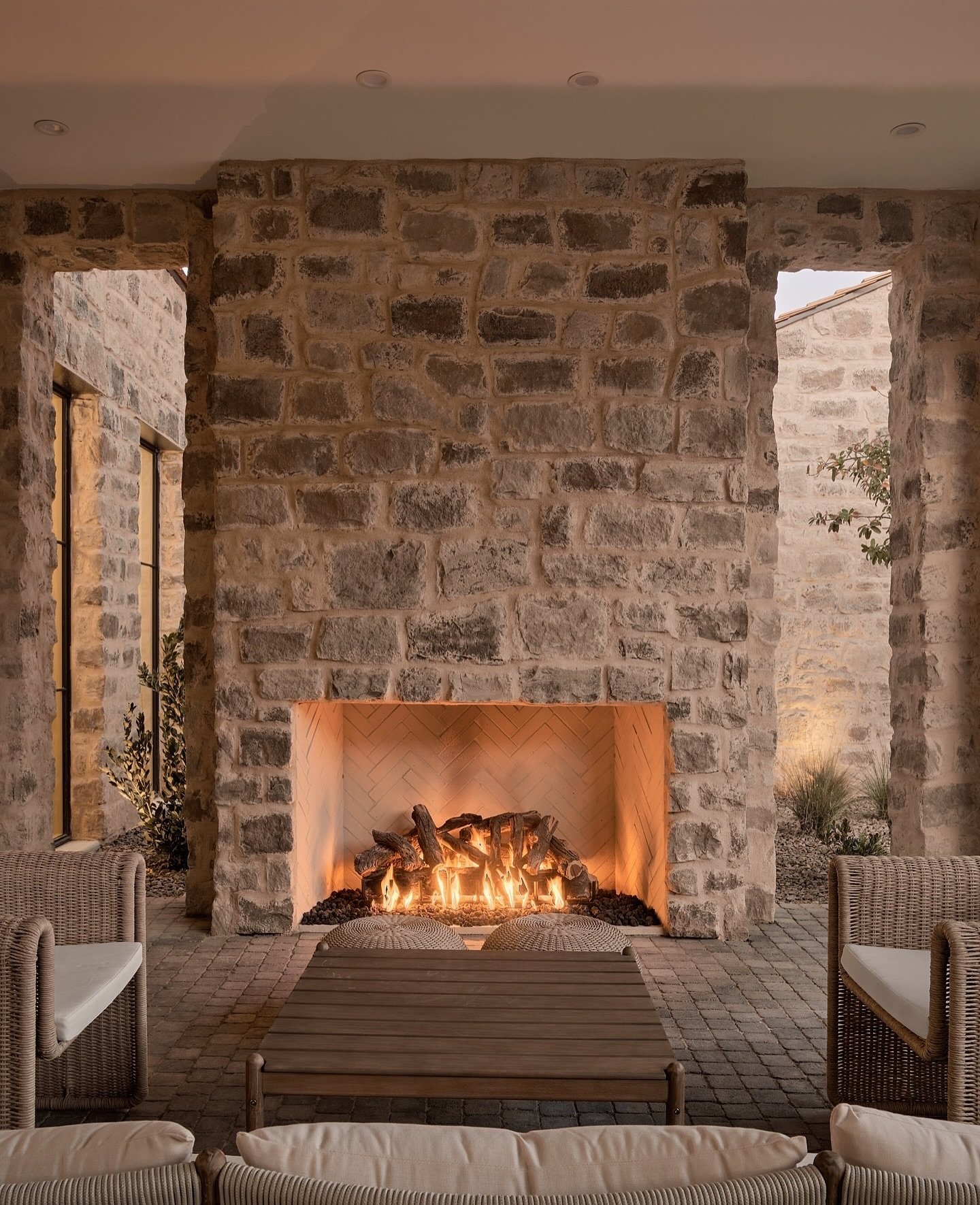 An outdoor patio fireplace built from large, rustic, light-toned rectangular cut stones, with a wide, glowing firebox featuring a chevron-patterned interior and wicker seating nearby.