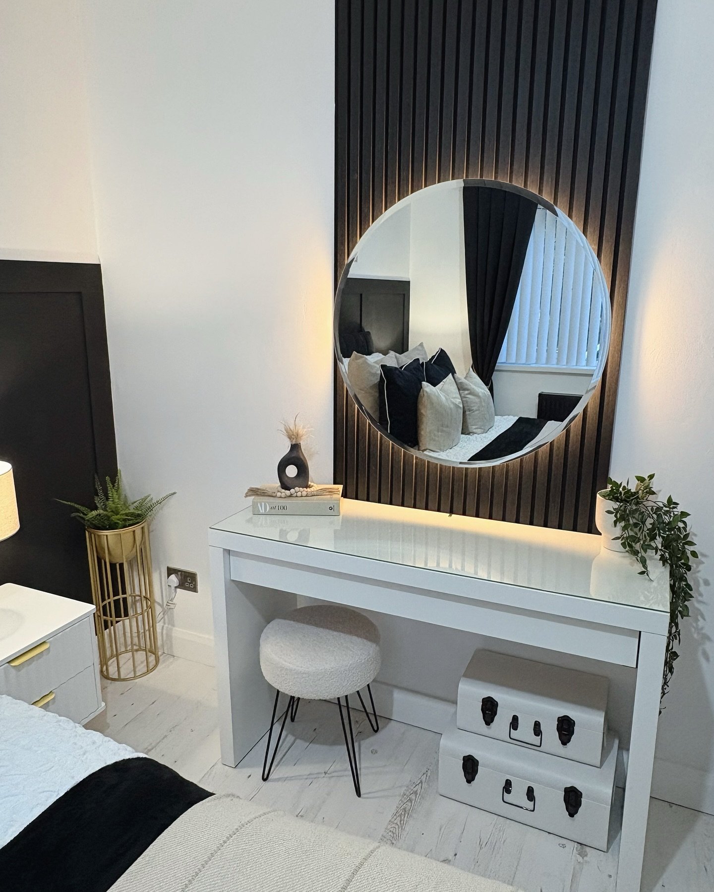 A modern white dressing table against a black slatted wall panel, featuring a large backlit round mirror, a white bouclé stool, and two stacked white storage trunks.