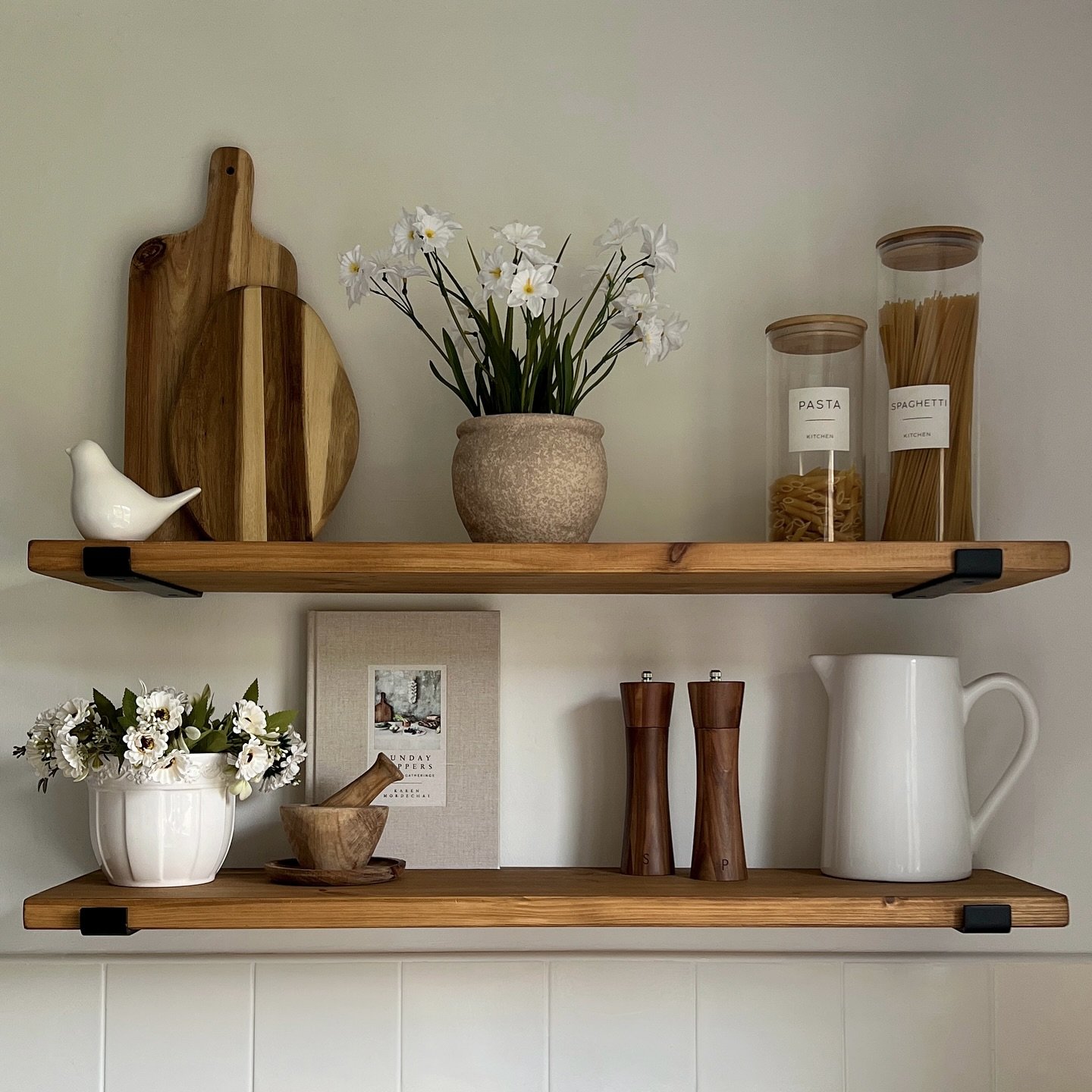 Two wooden kitchen shelves with black brackets, displaying pasta jars, cutting boards, and farmhouse decor.