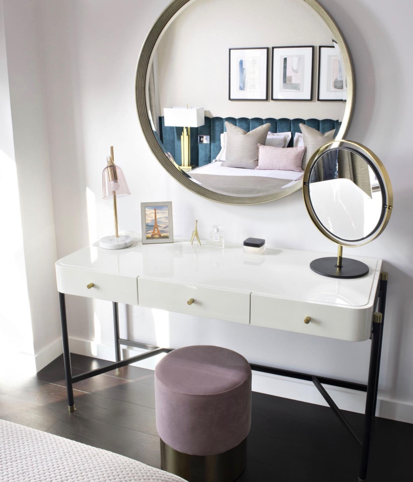 A sleek white high-gloss dressing table with three drawers and black metal legs, paired with a mauve velvet round stool and a large brass-framed round mirror.