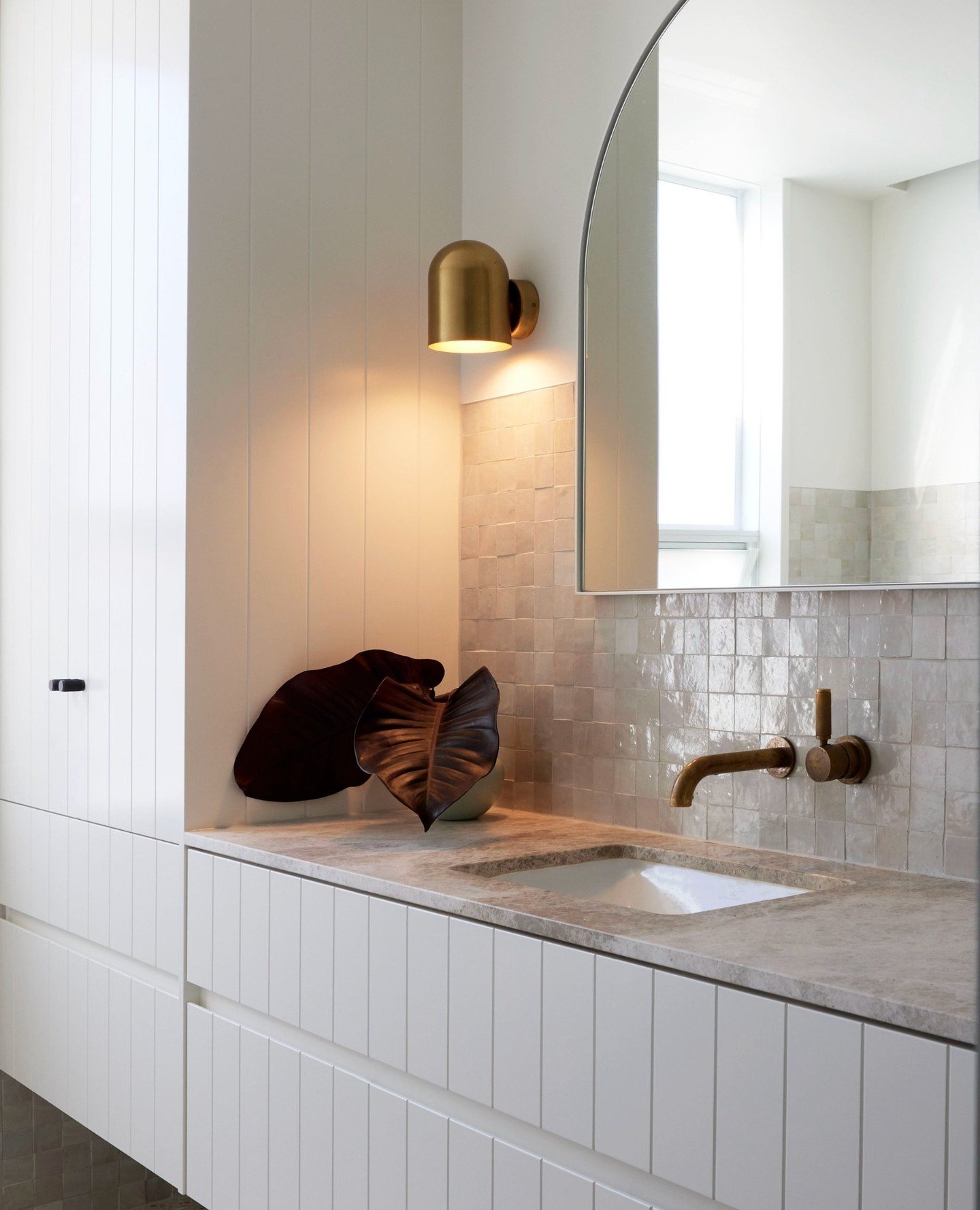 A single, rounded brass dome sconce casting light upward on a white paneled wall above a small-square, glossy tile backsplash.