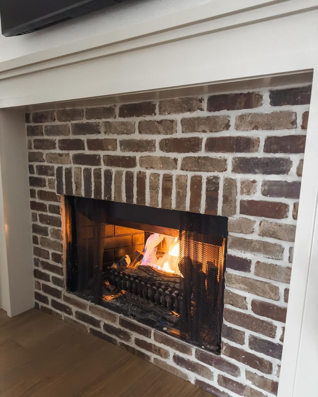 A close-up view of a fireplace with a traditional white wood mantel. The interior surround is faced with thin brick veneer in varied rustic colors and thick white mortar joints.