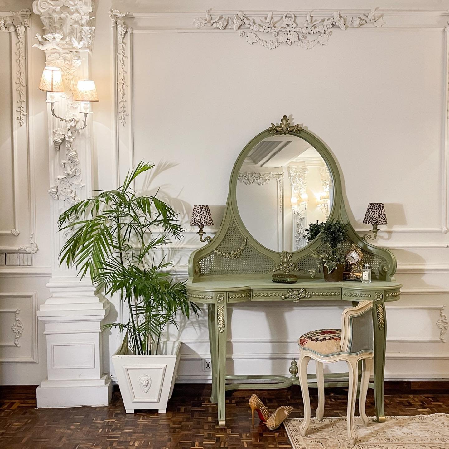 An ornate vintage-style dressing table in sage green with carved details and a cane back, featuring an oval mirror, two small lamps, and a decorative antique chair, set against an elaborately detailed white wall.