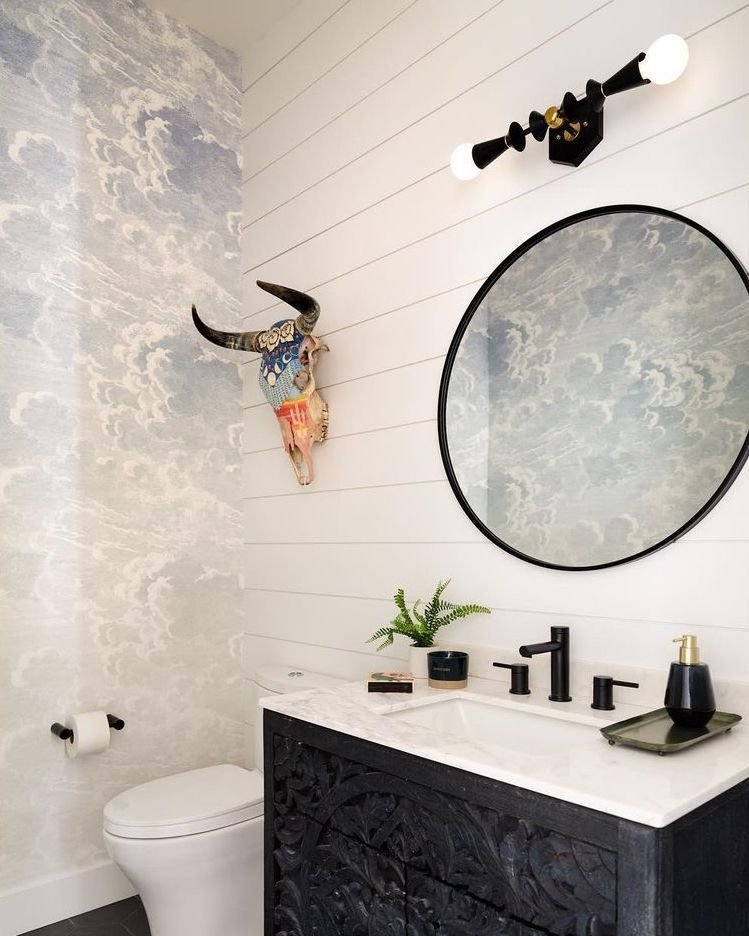 Horizontal black and brass sputnik-style sconce with exposed bulbs above a round black mirror and carved black vanity cabinet.