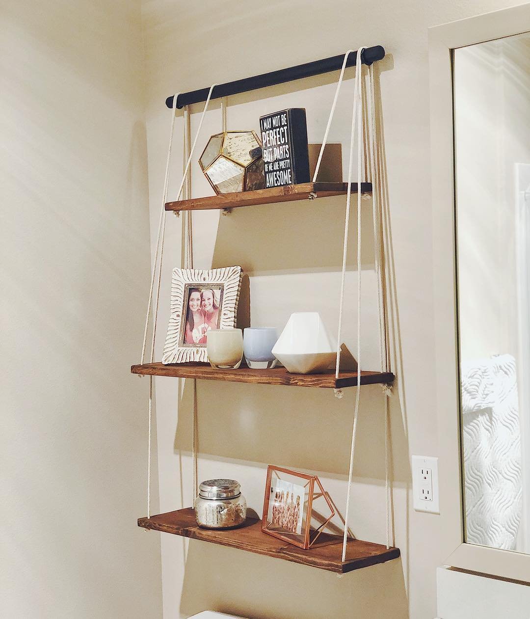A three-tiered wooden shelf unit hanging by white ropes from a black bar on a beige bathroom wall.