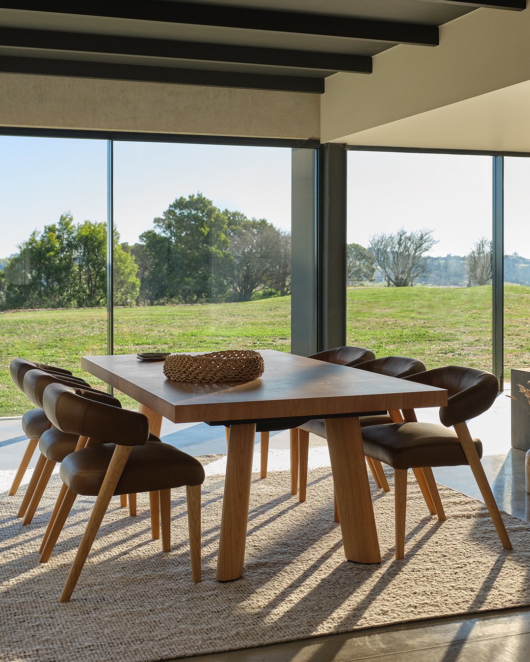 Wooden dining table with angled legs and brown leather chairs in front of a large window