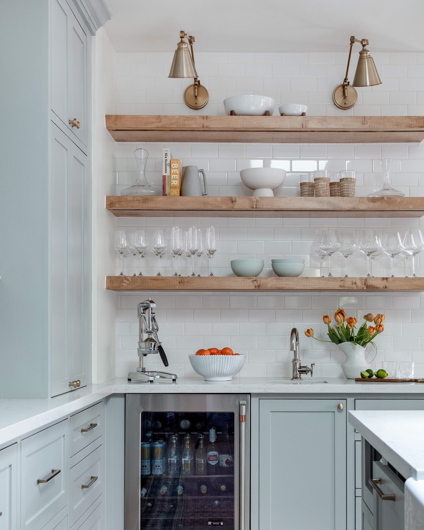 Three thick rustic wood floating shelves above a beverage bar sink and wine fridge, styled with glassware.