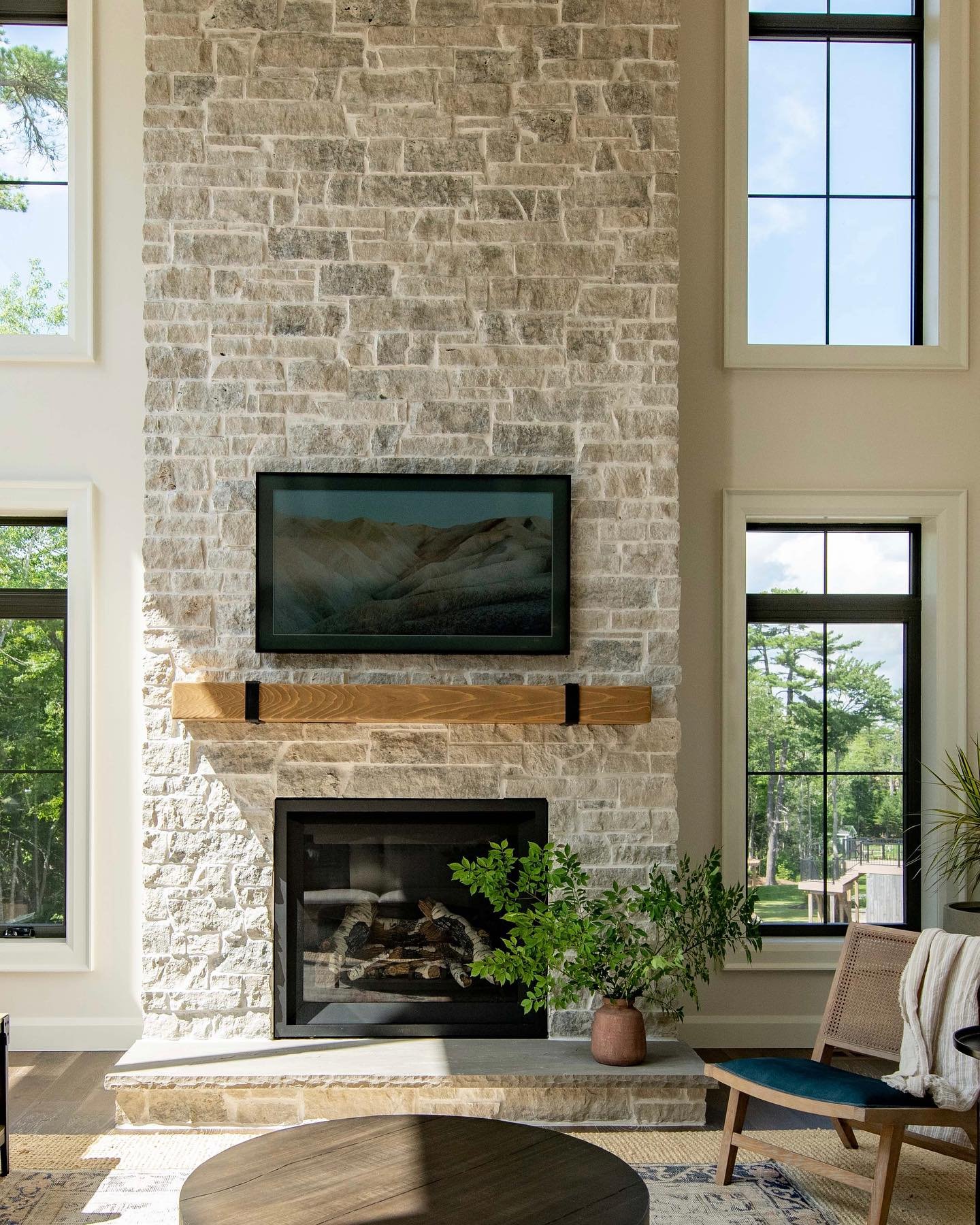 A tall, floor-to-ceiling stone fireplace made of light tan and cream-colored rectangular-cut stone, featuring a thick natural wood mantel supported by dark metal brackets, with a television mounted above.