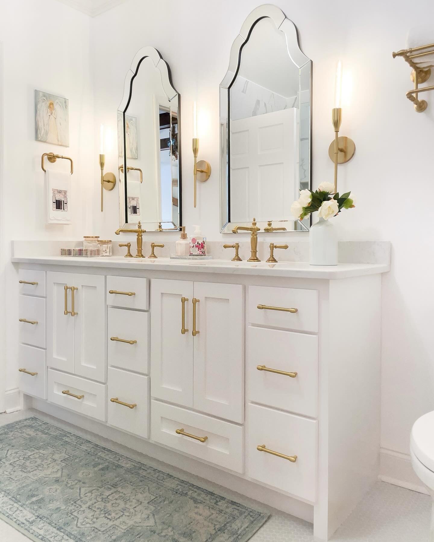 Symmetrical double vanity in white with gold hardware, featuring four tall, brass candle-style wall sconces flanking two arched, decorative mirrors.
