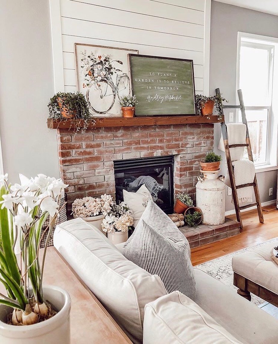 A cozy living room fireplace with natural red brick and a wood mantel. The area above the mantel is covered with white shiplap paneling.