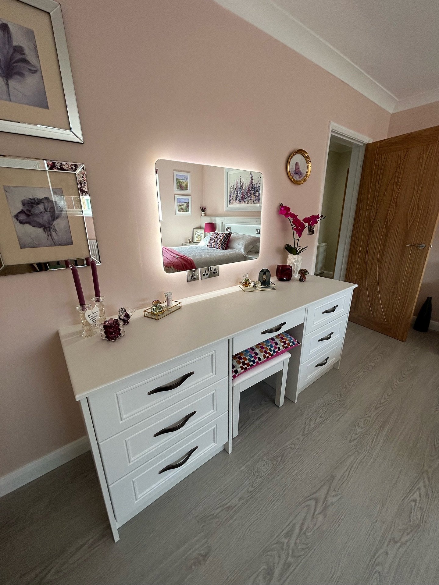 A wide, white dressing table and chest of drawers with eight drawers and black handles, featuring a central cutout for a stool, and a rectangular LED-backlit mirror mounted on a pink wall.