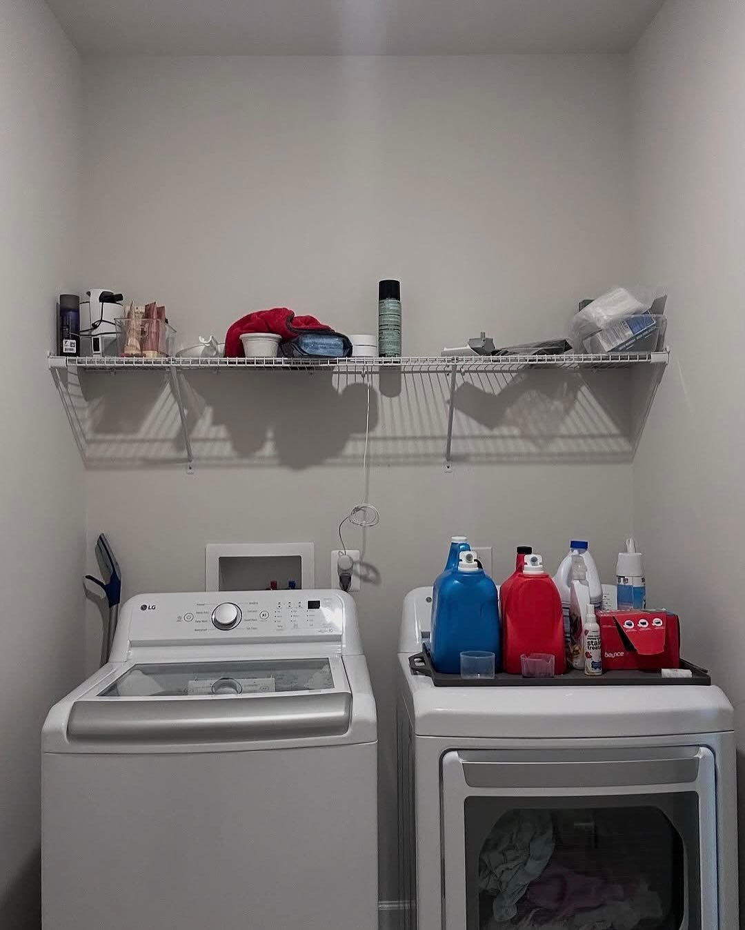 White wire laundry shelf holding miscellaneous cleaning supplies above a top-loading washer and dryer