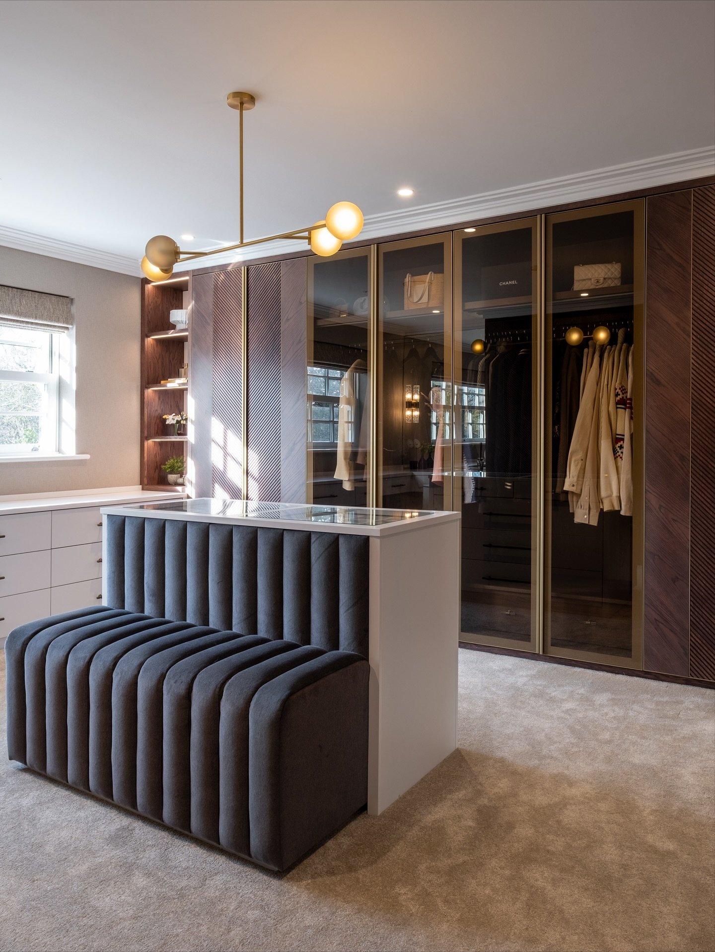 A contemporary dressing room with a dark wood and glass-fronted wardrobe, a unique central island featuring a ribbed grey velvet bench, and a modern gold linear chandelier.