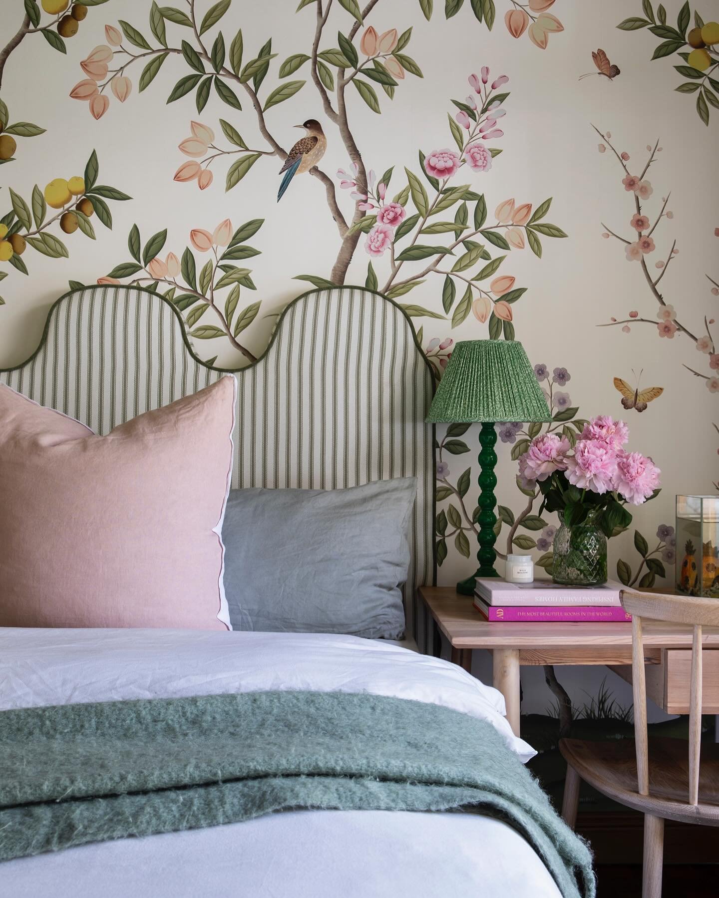 A close-up of a bed featuring a decorative Chinoiserie wallpaper with pink blossoms and green leaves, a striped green headboard, and a knitted green lamp.