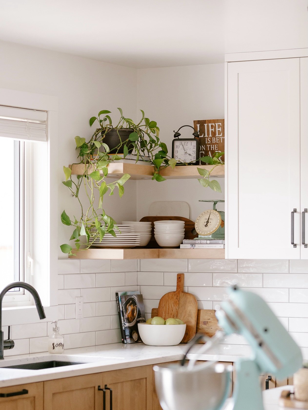 Two tiers of light wood L-shaped floating shelves holding plants and dishes next to white cabinets.