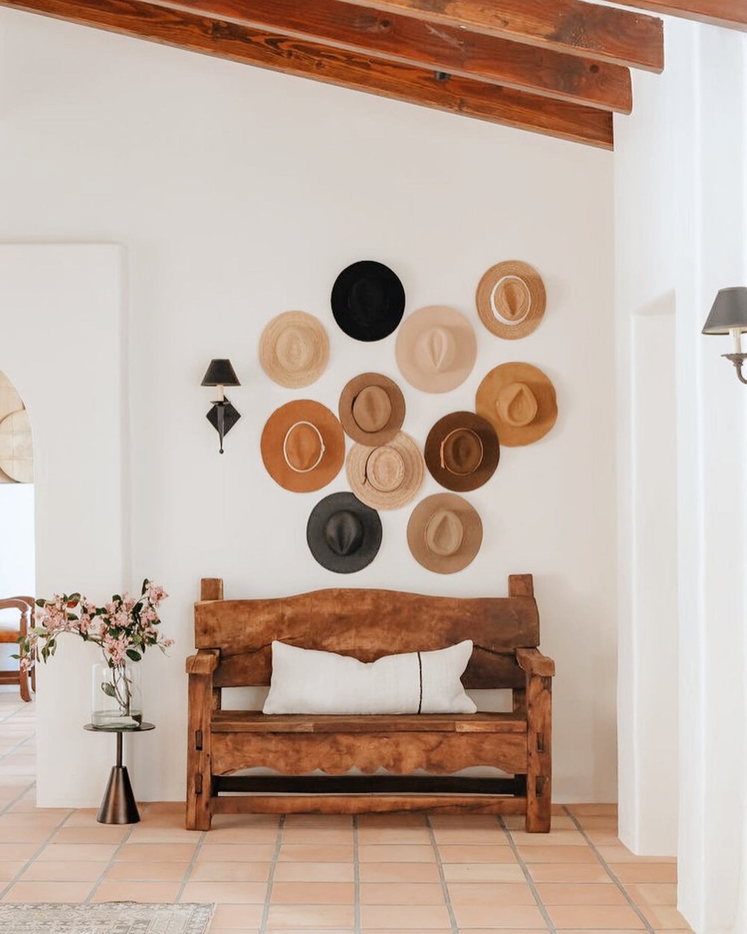 A white hallway wall features a collection of straw and felt hats arranged above a rustic wooden bench.