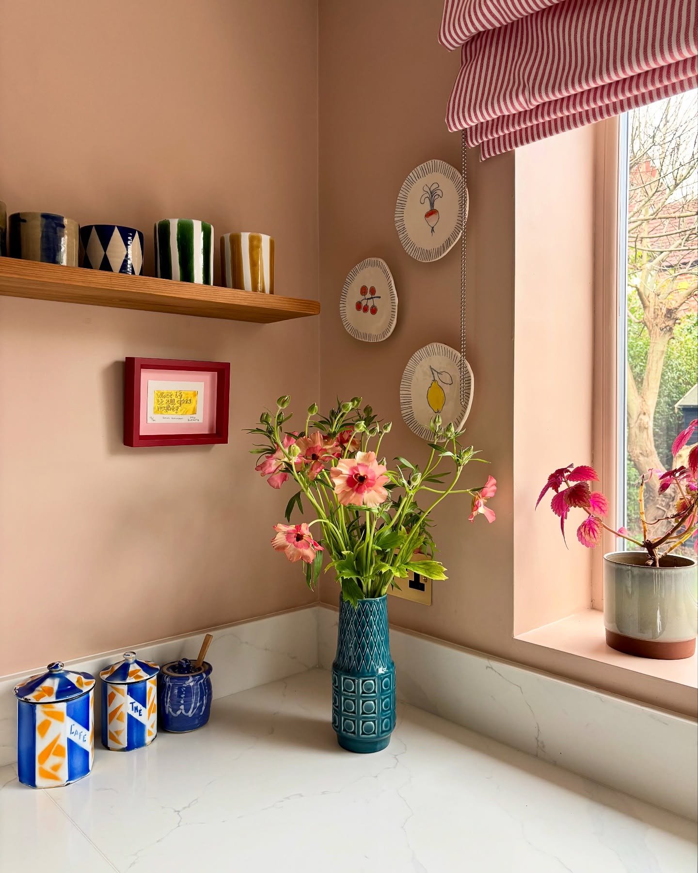 Pink kitchen corner featuring hanging vegetable plates and striped roman blind
