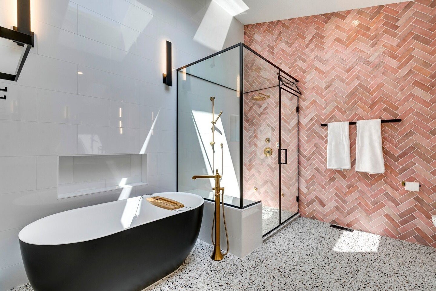 Modern bathroom with pink herringbone tile wall, terrazzo floor, and black freestanding tub.