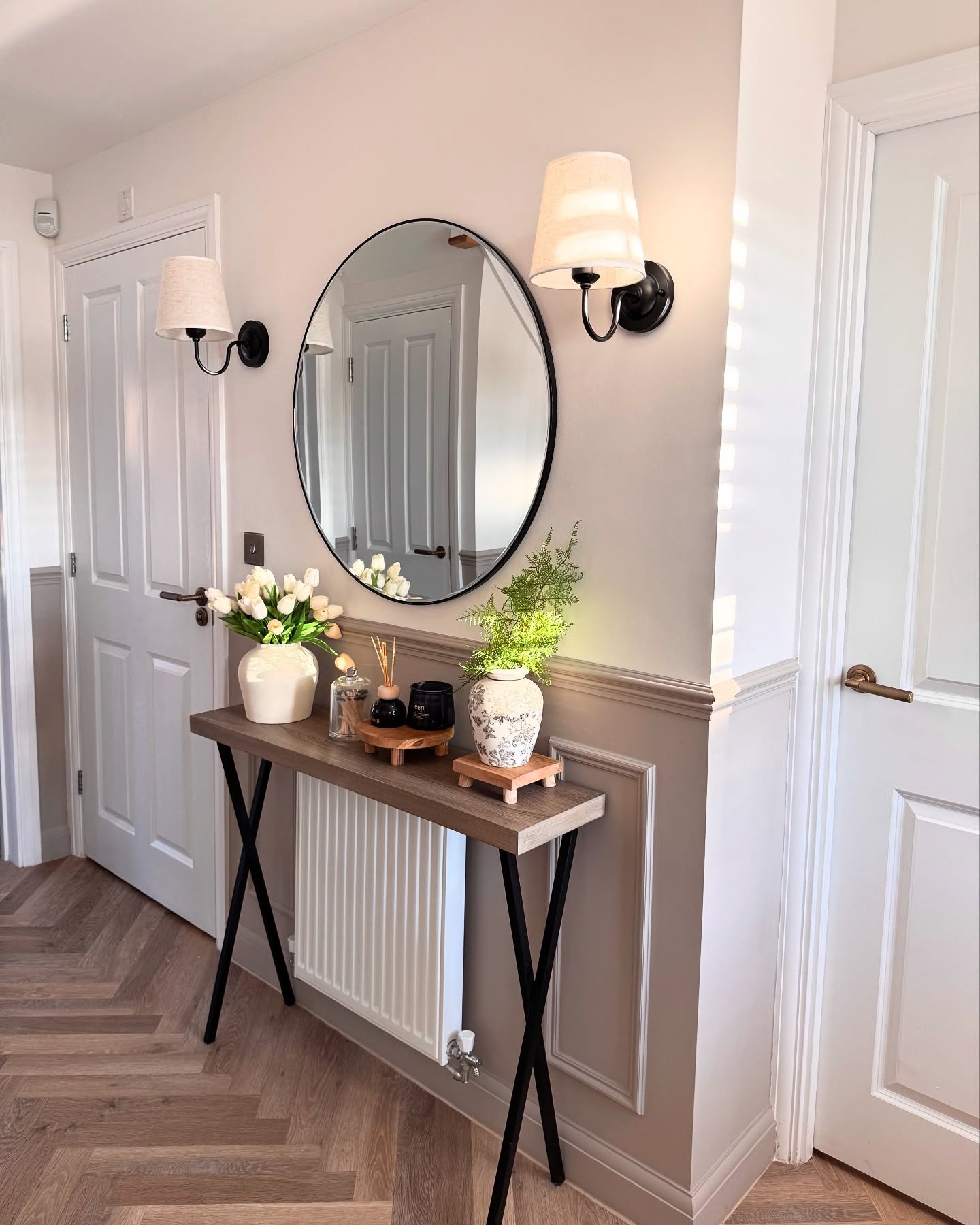 A hallway features grey wainscoting, a round mirror flanked by sconces, and a console table over a radiator with flowers.