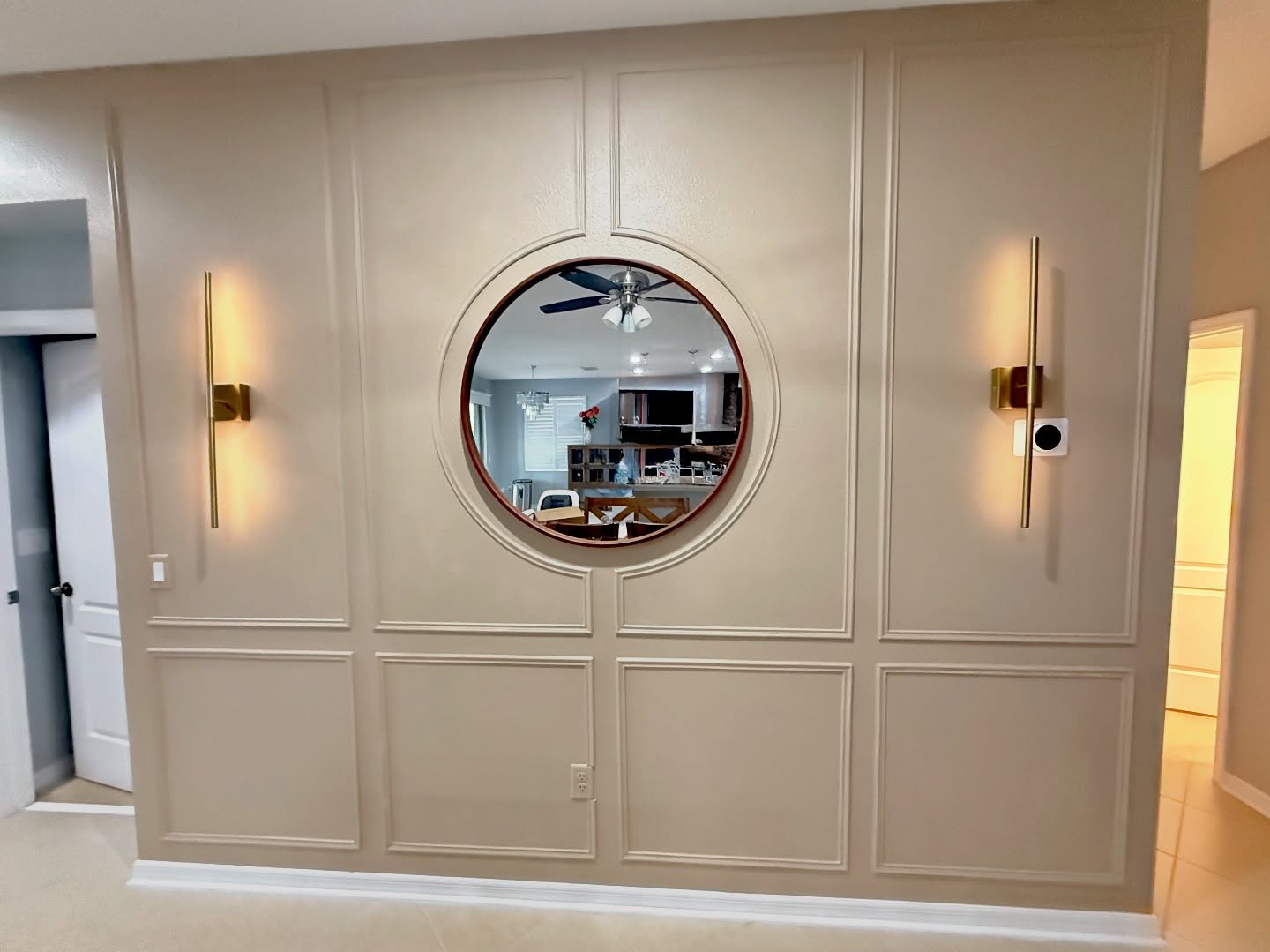 Beige entryway accent wall featuring picture frame molding, large round mirror, and gold wall sconces.